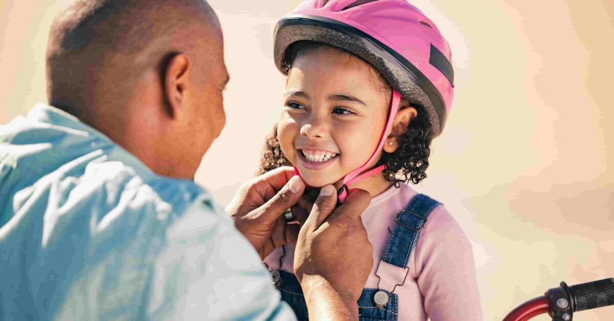 father putting a pink helmet on young daughter before she gets on bicycle